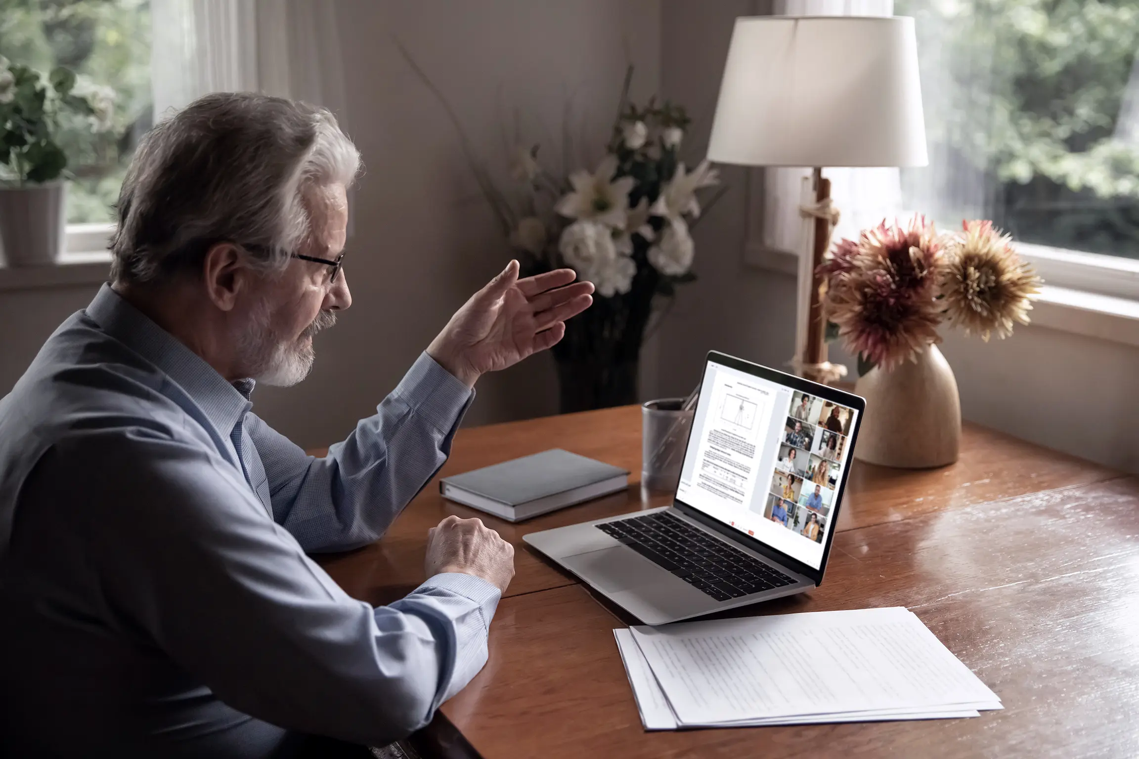 An educator in a cozy home office setting gesturing during a remote learning session. Visible on his laptop screen are a grid of online participants, and a lecture document that is being streamed to the meeting.