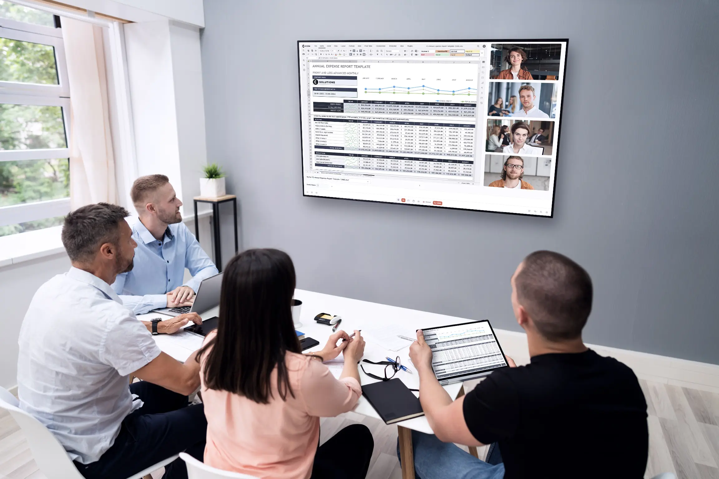 A team of professionals sitting at a conference table, engaged in a video call with remote participants displayed on a large monitor. A detailed spreadsheet is currently being screen shared.